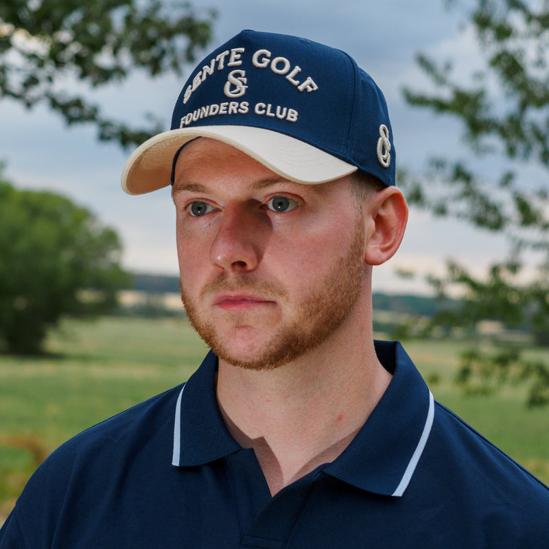 A man wearing a navy blue polo shirt and matching navy blue cap with 'SENTE GOLF SG FOUNDERS CLUB' embroidered in white, photographed outdoors against a blurred golf course background.