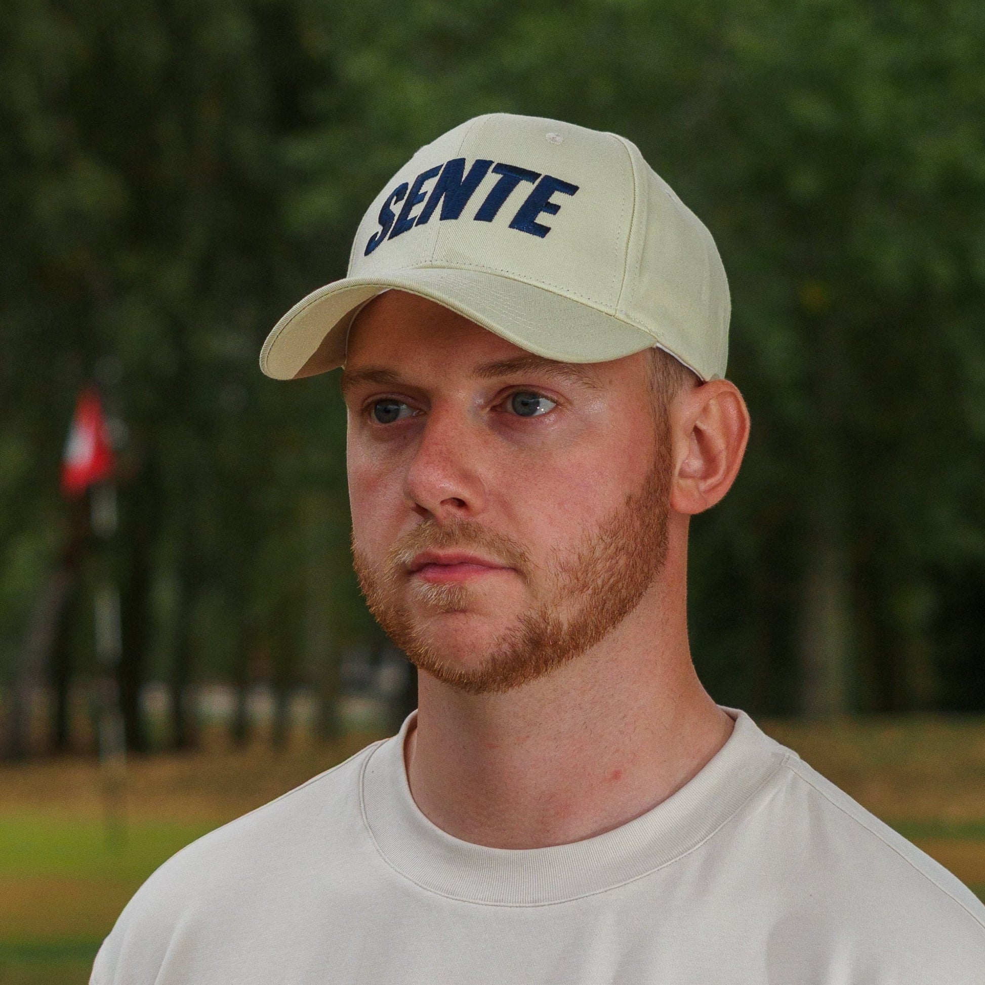 A man wearing a beige SENTE baseball cap and a matching beige crewneck t-shirt with dark green side panels, standing outdoors on a grassy field with trees in the background.