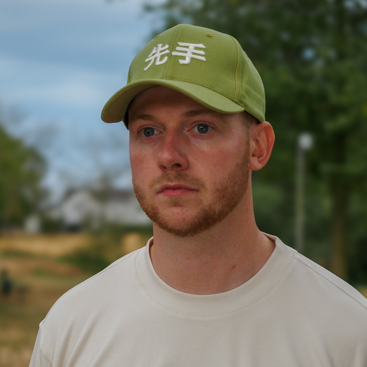 A man wearing a light green baseball cap with white Japanese characters and a cream-colored t-shirt with the brand name 'SENTE' printed on the chest, photographed outdoors against a blurred background of trees and sky.