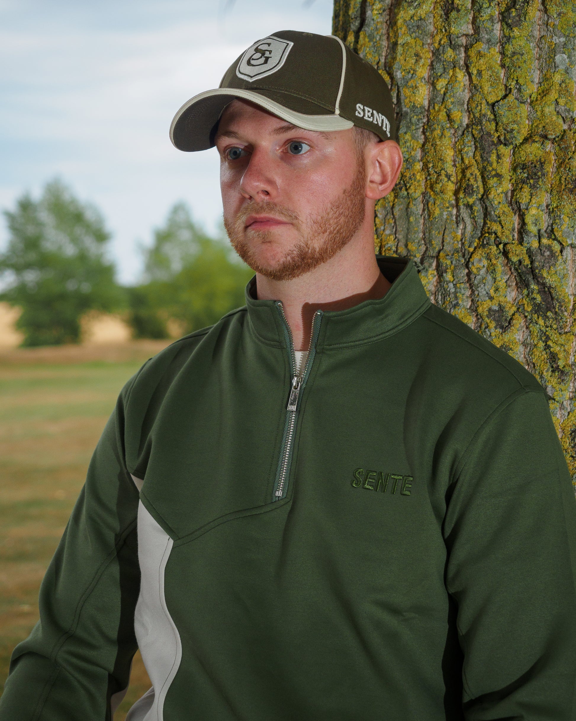 A man wearing a green SENTE quarter-zip fleece pullover and a matching green cap, standing outdoors next to a tree trunk.