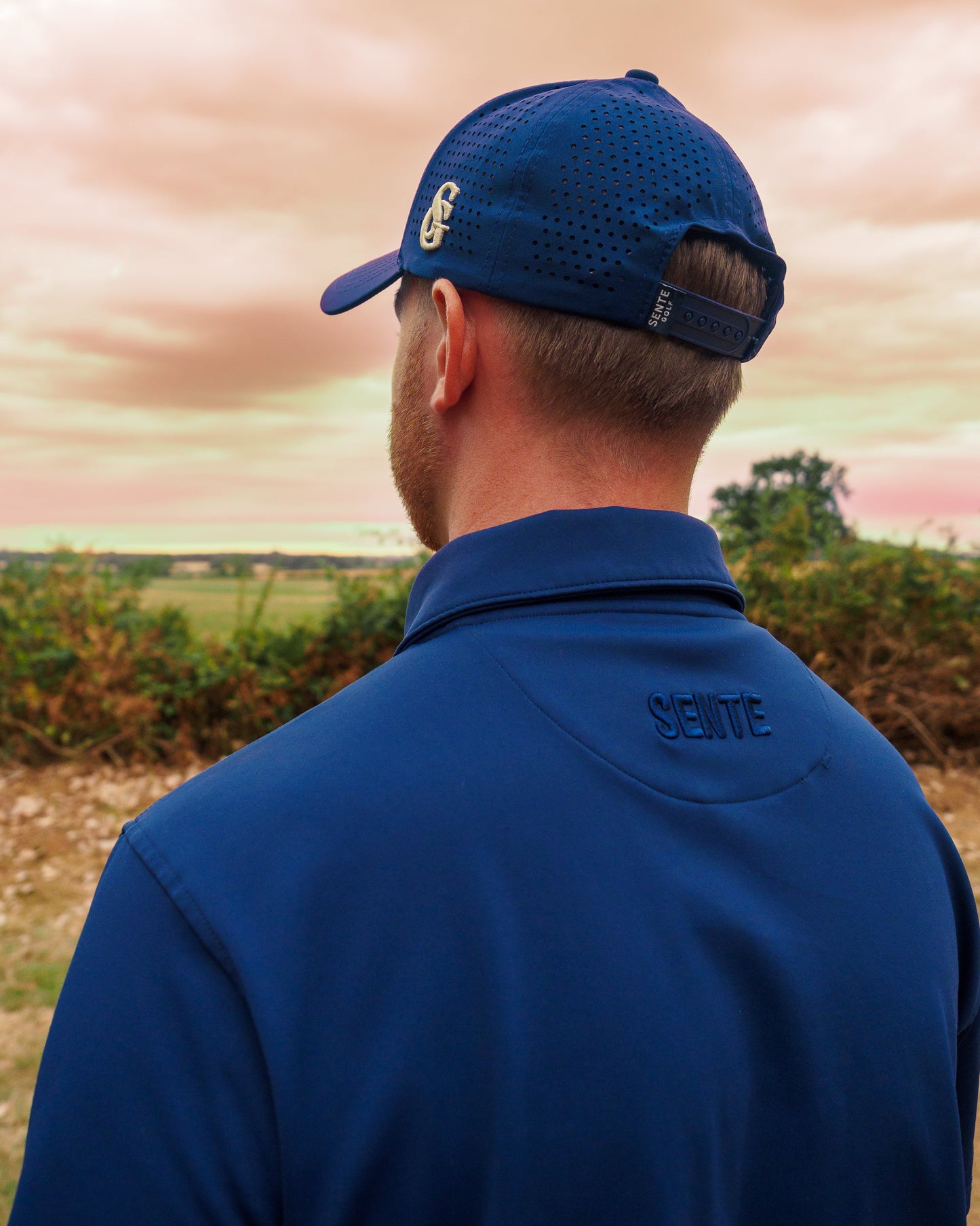 A man viewed from behind wearing a navy blue polo shirt and matching navy blue perforated baseball cap, standing outdoors against a soft-focus landscape and sky.