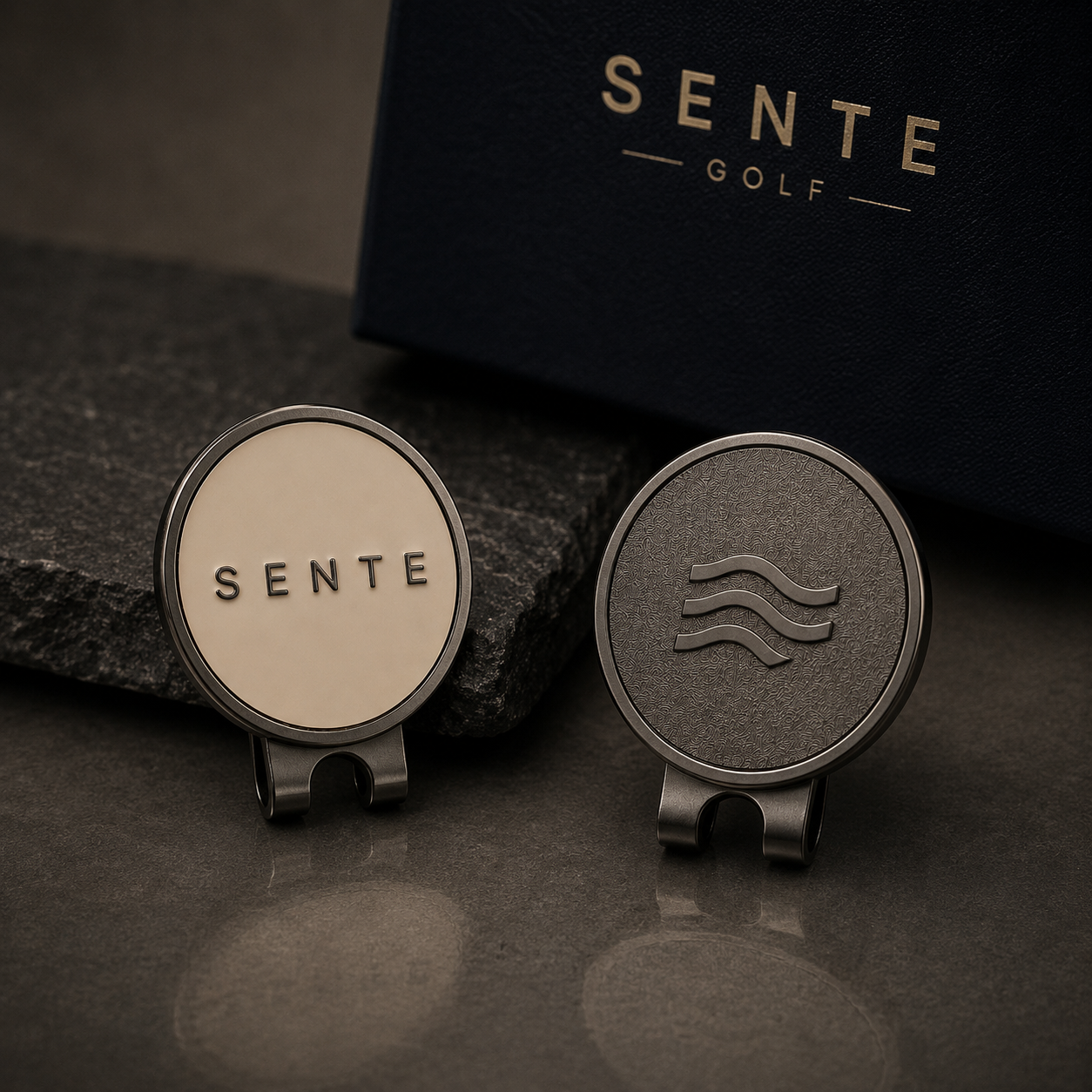 SENTE Ball Marker, golf accessories displayed on a dark surface, with a dark navy SENTE GOLF box in the background. One piece has a cream face with black 'SENTE' lettering, and the other has a textured dark gray face with a wave logo.