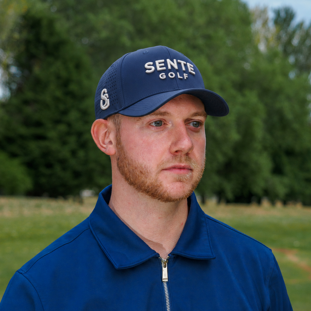 A man wearing a navy blue Sente Golf baseball cap and a matching navy blue polo shirt with a white color-block panel, standing outdoors on a golf course with trees in the background.