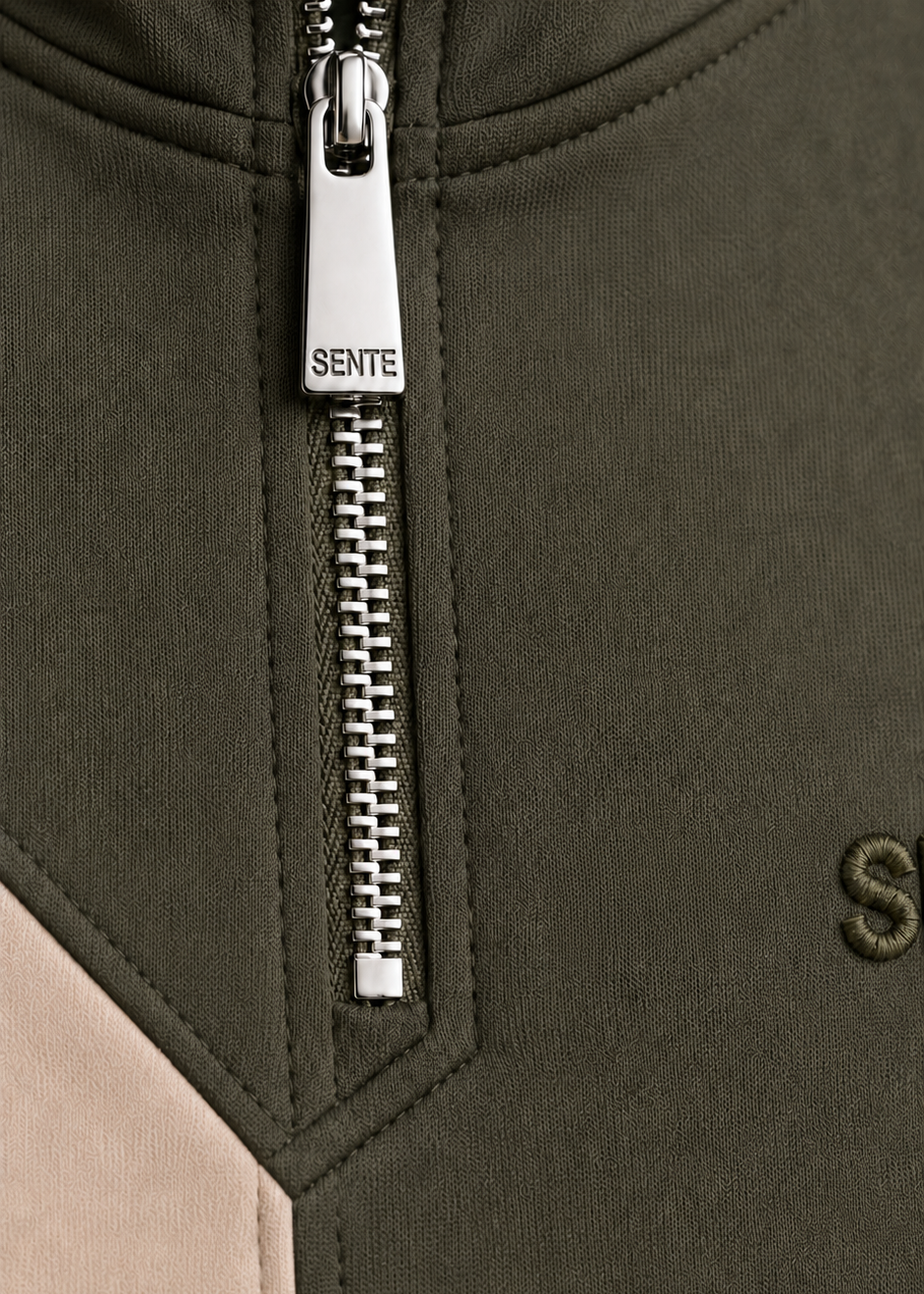 Close-up of a color-blocked zip-up jacket by SENTE, featuring an olive green body, a beige color-block panel on the left, and a silver metal zipper pull with the brand name 'SENTE' embroidered on it.