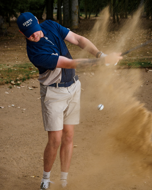 A golfer in a blue polo shirt, beige shorts, and a blue cap swings a club in a sand bunker, kicking up a large cloud of sand. Trees and grass are visible in the background.