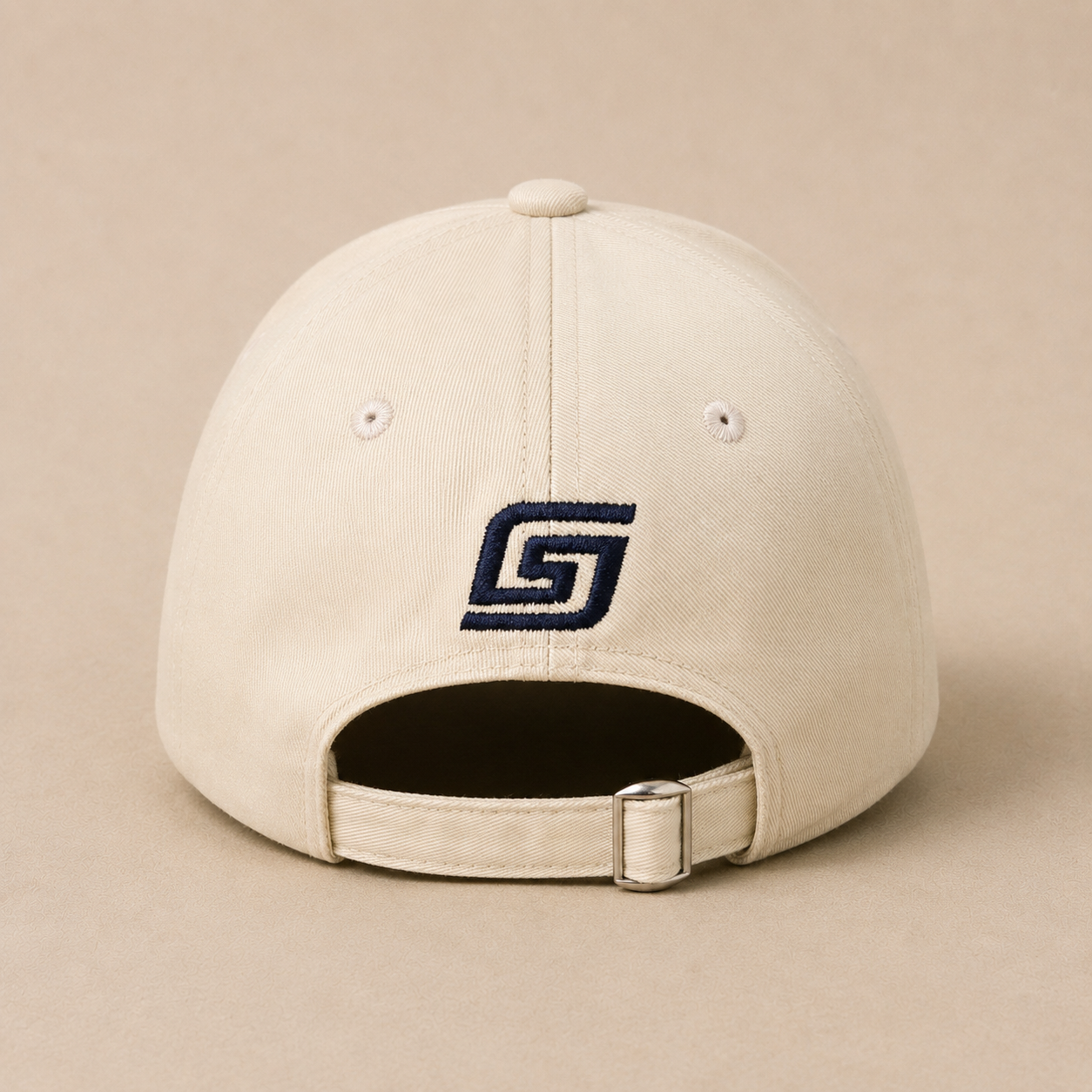 A cream-colored baseball cap shown from the back, featuring a navy blue embroidered stylized 'G' logo on the center back and an adjustable strap with a silver metal buckle, set against a solid beige background.